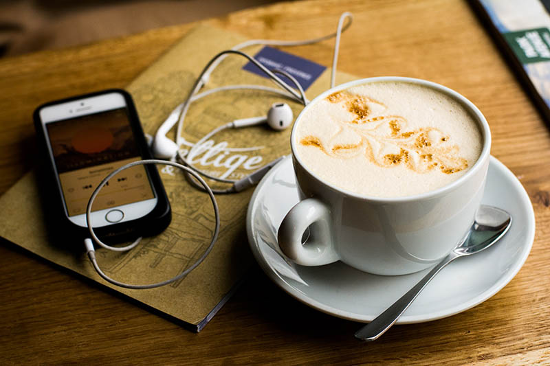 Photo of a phone with headphones tuned to creative business podcasts next to a cappuccino sitting on a table.